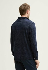 Tmavomodrá polo košeľa s dlhým rukávom a falošným golierom, s melírovanou textúrou a rovným lemovaním, predvedená na modeli otočenom chrbtom.