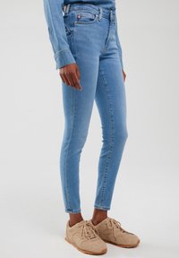 Osoba odjevena u svijetloplave traperice uskog kroja, bež tenisice s vezicama i odgovarajuću svijetloplavu traper košulju, stojeći na bijeloj podlozi.