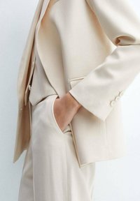 Beige oblek na míru s dobře padnoucí sakem s klopou, kapsou a perlovými detaily na manžetách. Hladká, lehká tkanina.