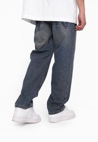 Jeans in denim scuro con una vestibilità ampia, caratterizzati da leggeri sbiadimenti e dettagli di cucitura. Abbinati a sneakers bianche, mostrando superfici texturizzate.