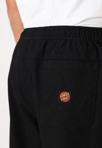 Sorte joggebukser med elastisk talje, som har en baglomme med et rundt "Santa Cruz" logo i rød og grøn. Glat tekstur.