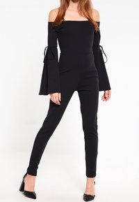 Svart off-shoulder jumpsuit med långa trumpetärmar. Har knytaccent vid armbågarna och en slim fit-design genom benen.