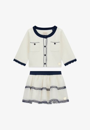 Weißer Cardigan mit marineblauer Einfassung, Knopfverschluss, zwei Vorderentaschen und gewellten Kanten, kombiniert mit einem mehrlagigen Tüllrock mit Spitzenakzenten.