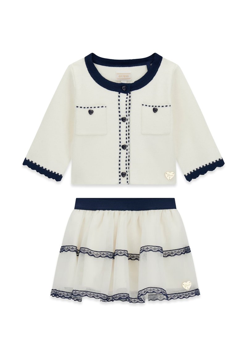 Cardigan blanc avec bordure marine, fermeture à boutons, deux poches frontales et bords festonnés, associé à une jupe en tulle à couches avec des accents en dentelle.