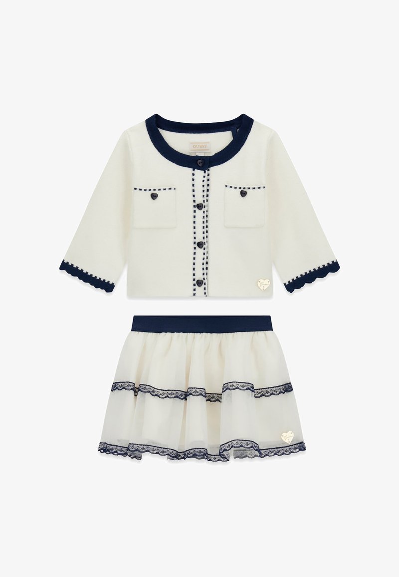 Cardigan blanc avec bordure marine, fermeture à boutons, deux poches frontales et bords festonnés, associé à une jupe en tulle à couches avec des accents en dentelle.