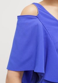 Blå topp med off-shoulder design, flödande ärmar och glittrande detaljer på axelbanden. Tyget verkar glatt och lätt.