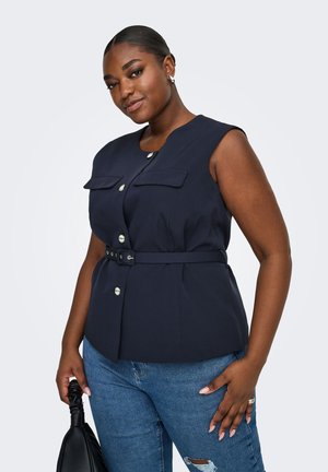 Femme portant un haut sans manches bleu marine avec des boutons et une ceinture, jean bleu déchiré, tenant un sac à main noir sur un fond clair uni.