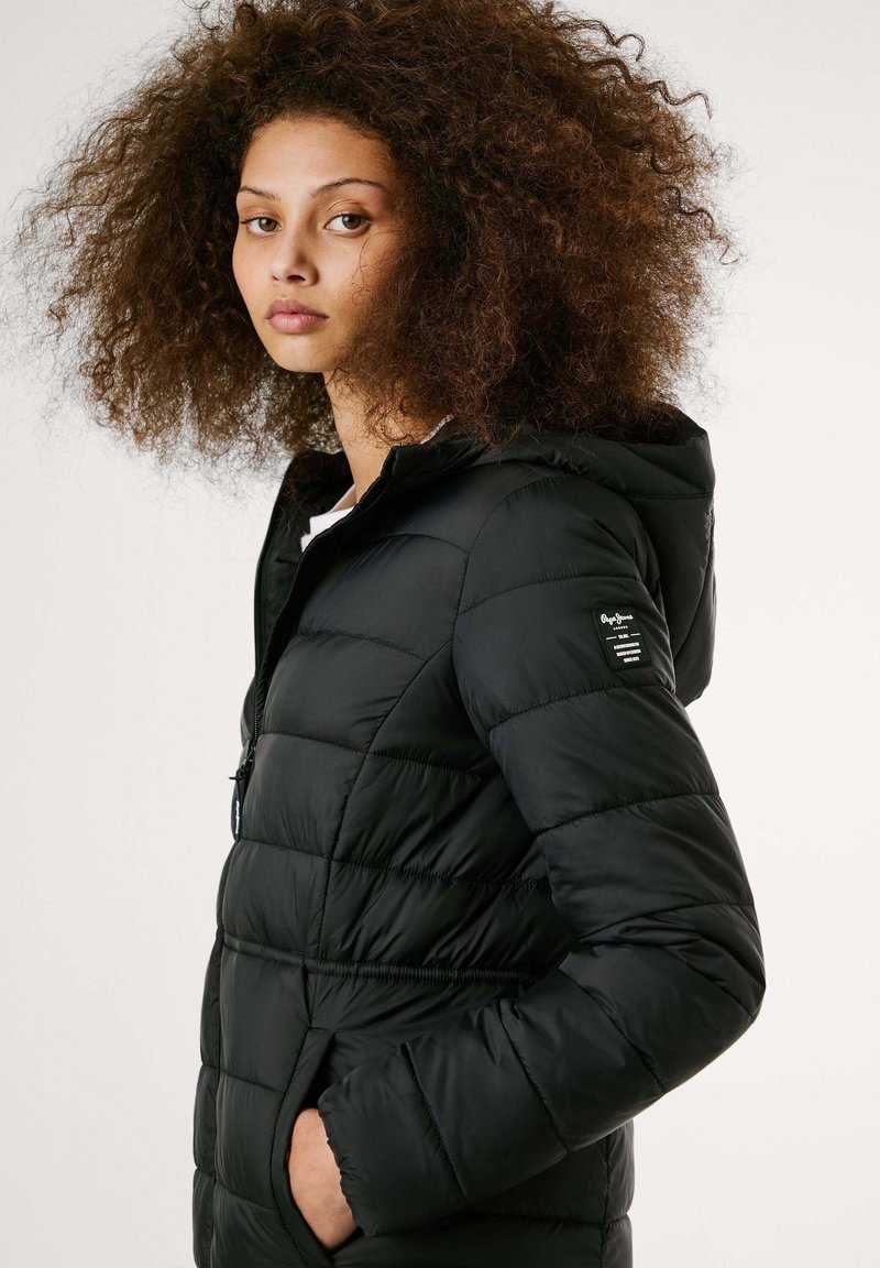 Giacca nera imbottita con cappuccio, caratterizzata da sezioni trapuntate, una zip anteriore e una patch con logo sulla manica sinistra. Tessuto liscio.