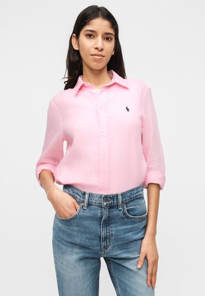 Femme aux cheveux foncés portant une chemise rose pâle à boutons avec les manches retroussées et un jean bleu, debout devant un fond blanc uni.