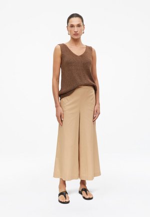 Femme debout, portant un haut sans manches marron texturé, un pantalon large beige, des sandales noires et des boucles d’oreilles créoles dorées, sur fond blanc.