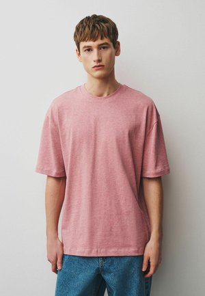 Junger Mann mit kurzem braunem Haar, der ein locker sitzendes, rosa kurzärmeliges Hemd und blaue Jeans trägt, steht vor einem schlichten hellen Hintergrund.