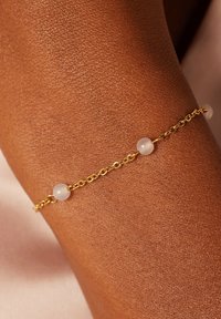 Goldene Kettenarmband mit drei durchsichtigen Perlen, vor strukturiertem Hauthintergrund. Verfügt über ein zartes Design und erscheint leicht.