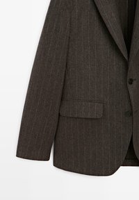 Brauner Blazer mit Nadelstreifen, mit Reverskragen, Ein-Knopf-Verschluss und einer Tasche auf der linken Seite. Der Stoff hat eine strukturierte Oberfläche.