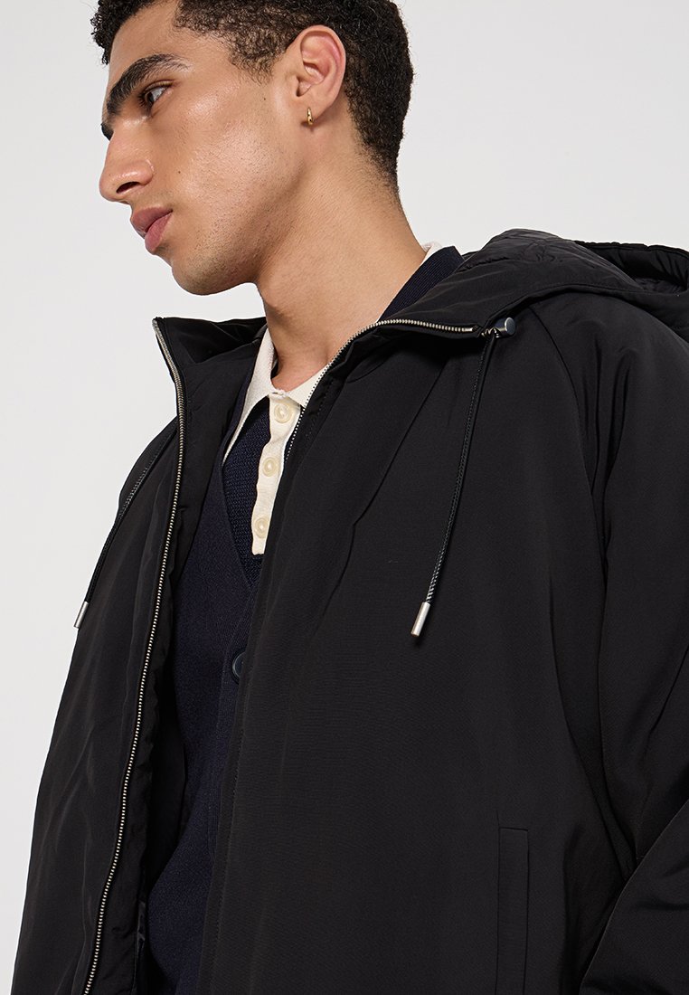 Zwarte hoodie met een rits aan de voorkant, voorzien van een trekkoordsluiting, textuurstof en zijzakken, gelaagd over een marineblauw trui.
