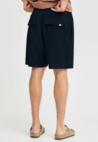 Navy blue shorts with textured fabric, two back flap pockets, and a small logo on the waistband. Worn with beige sandals.
