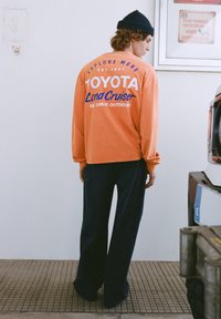 Oranžová košeľa s dlhým rukávom s nápisom „TOYOTA Land Cruiser“ modrou farbou. Kombinovaná s tmavomodrými širokými nohavicami. Na hlave vlnená čiapka.