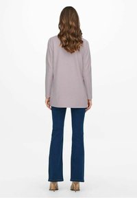 Top viola chiaro a maniche lunghe, leggermente più lungo sul retro, abbinato a jeans a zampa d'elefante blu e scarpe con tacco beige. Texture liscia.