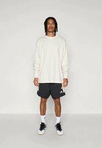 Man standing wearing oversized white long-sleeve shirt, black Adidas shorts with logo, white socks, and white and black sneakers.