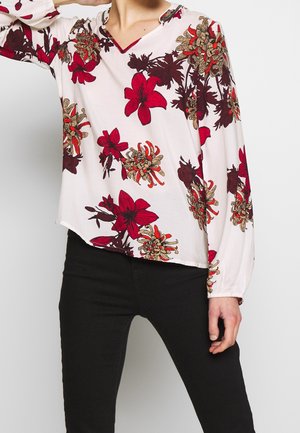 Person iført hvid bluse med lange ærmer med rød og brun blomsterprint og sorte bukser mod ensfarvet baggrund.