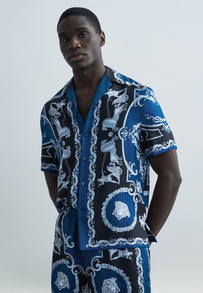 Chemise à manches courtes en tissu bleu et noir avec des motifs argentés complexes, comprenant des motifs de lion et de chérubin, texture lisse et fermeture par boutons.