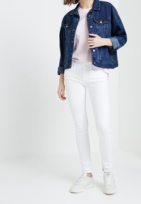 Mujer con chaqueta de mezclilla azul oscuro, camiseta rosa clara, jeans blancos ajustados y zapatillas blancas de pie contra un fondo blanco liso.