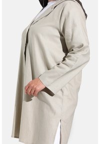 Veste chemise beige à manches longues avec un col pointu, des boutons à l'avant, des fentes latérales et une texture lisse.