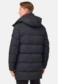 Man som bär en lång svart pufferjacka med huva, sedd bakifrån, stående mot en enkel vit bakgrund.