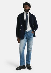 Blazer marinero de doble botonadura sobre una camisa de rayas blancas y corbata de lunares, combinada con jeans de color azul claro y zapatos negros con cordones.