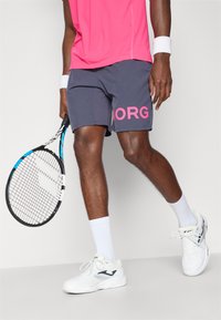 Ανδρική αθλητική ενδυμασία τένις με ροζ πουκάμισο και γκρι σορτσάκι με το "BORG" σε φωτεινό ροζ. Λευκά παπούτσια με σκούρες λεπτομέρειες και λευκές κάλτσες.