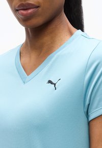 Κλείσιμο πλάνου ενός ατόμου που φοράει ανοιχτό μπλε μπλουζάκι V-λαιμόκοψη με μαύρο λογότυπο Puma στο στήθος.