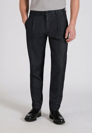 Mężczyzna ubrany w czarne plisowane spodnie z bocznymi kieszeniami i czarne skórzane mokasyny, stojący na jednolitym tle.