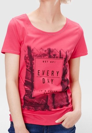 Kobieta nosząca jasno różową koszulkę z czarną grafiką i napisem „WSTAŃ! KAŻDY DZIEŃ TO NOWY POCZĄTEK”, w połączeniu z niebieskimi dżinsami.