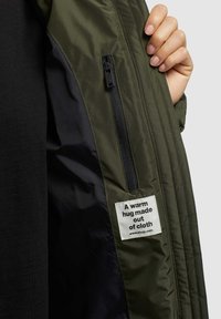 Chaqueta verde oliva con una textura suave, bolsillo interior con cremallera y una etiqueta que dice "Un abrazo cálido hecho de tela." Forro negro visible.
