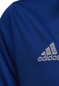Maglietta sportiva blu realizzata in tessuto morbido, presenta un logo Adidas ricamato in argento sul lato destro del petto, con texture e cuciture sottili.
