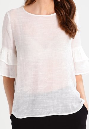 Kobieta w półprzezroczystej białej bluzce z falbanami na rękawach do połowy przedramion i czarnych spodniach, z rękami w kieszeniach.