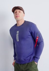 Felpa viola con scollatura a girocollo, dotata di un logo frontale e di una tasca con zip rossa sulla manica. Realizzata in tessuto morbido.