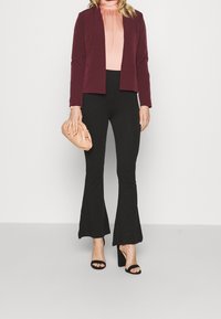 Blazer bordeaux su una blusa rosa chiaro, abbinato a pantaloni neri svasati. Accessoriato con una borsa a mano beige e sandali neri con tacco alto.