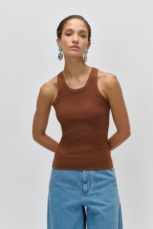 Žena s pletenými vlasy, která má stříbrné kruhové náušnice, hnědý žebrovaný top bez rukávů a světle modré džíny s vysokým pasem, stojí s rukama za zády.