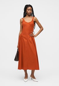 Γυναίκα με μακριά πλεξούδες που φοράει πορτοκαλί αμάνικο μίντι φόρεμα, κρατώντας σκούρα τσάντα και φορώντας λευκά μυτερά τακούνια, στέκεται μπροστά σε λευκό φόντο.