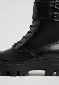 Črni usnjeni gležnjarji z vezalkami spredaj, zaponko okoli gležnja in grobo profiliranim podplatom za dober oprijem.