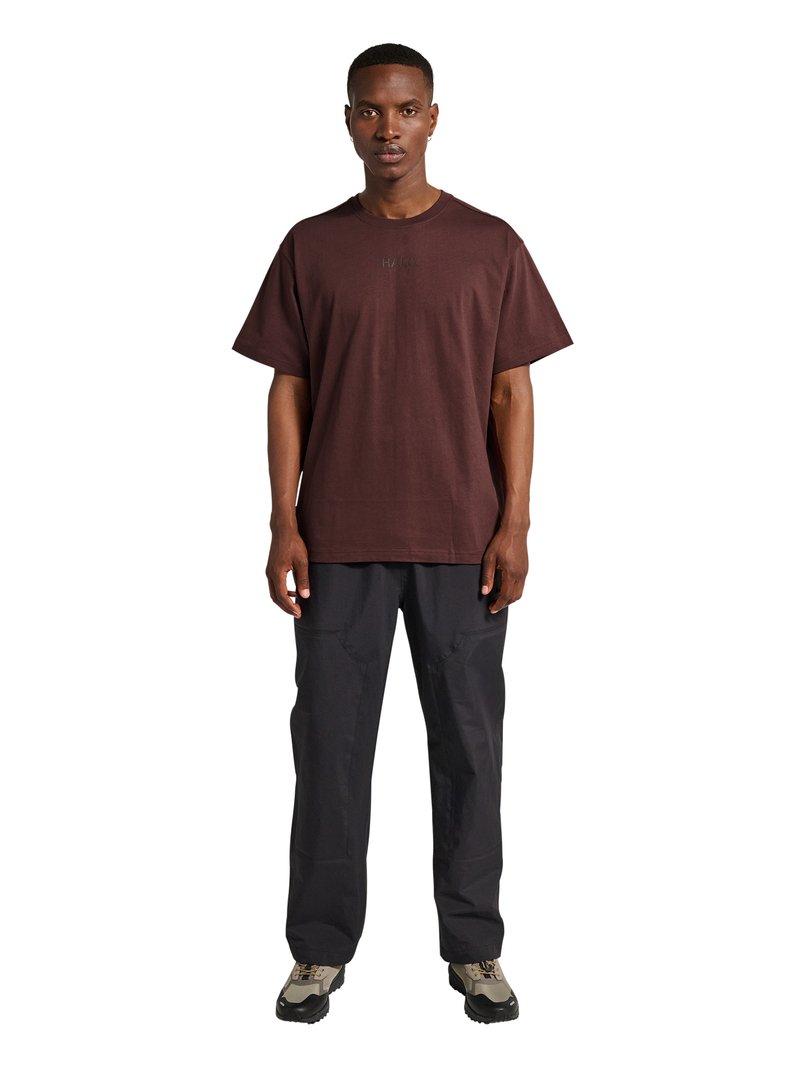 Mand stående iført en mørkebrun oversized t-shirt, sorte cargobukser og beige og sorte vandresko på hvid baggrund.