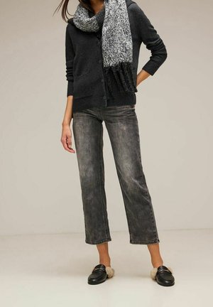 Henkilö pukeutuneena tummaan napitettuun neuletakkiin, harmaaseen ja mustaan huiviin, kuluneisiin mustiin lyhennettyihin farkkuihin ja mustiin töppösaappaisiin, joissa on karvavuori.