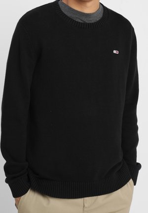 Mand iført sort crewneck-trøje med lille rektangulært rødt, hvidt og blåt logo på venstre bryst, grå skjorte under og beige bukser.