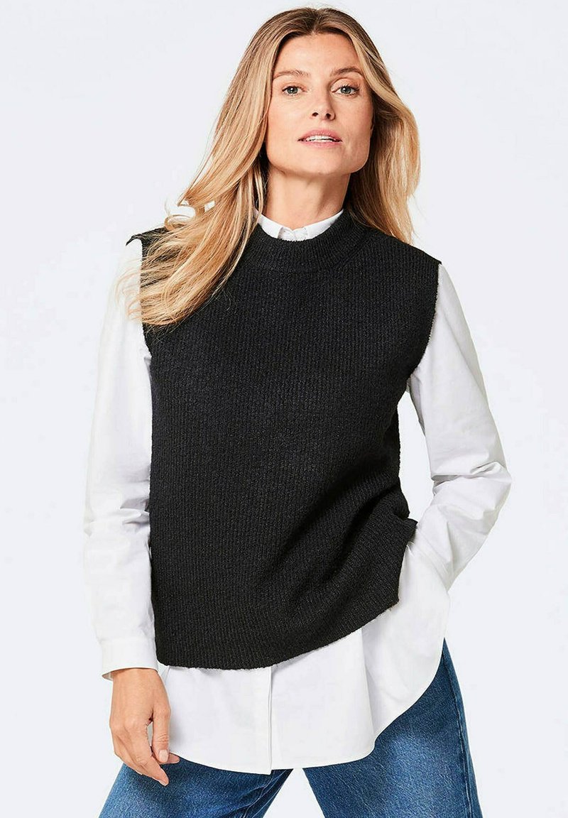 Schwarzer ärmelloser Pullover mit strukturiertem Strickdesign, getragen über einem weißen Hemd mit Kragen. Hat einen runden Ausschnitt und eine lockere Passform.