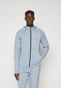 Veste à capuche bleu clair avec fermeture éclair, fabriquée en matériau doux. Équipée d'une fermeture éclair noire et d'un logo sur la poitrine, avec une coupe ajustée et une texture lisse.