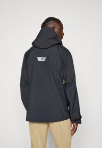 Zwarte waterdichte jas met een capuchon, voorzien van een zilveren logo patch op de achterkant en contrasterende groene ritsaccenten op de mouwen.