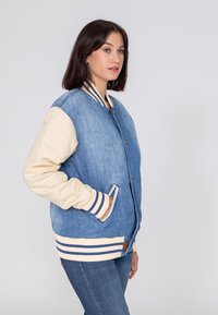 Veste en denim avec des manches crèmes, des poignets côtelés et un bas. Présente un devant à boutons, des poches latérales et des accents rayés. Couleur bleu clair.