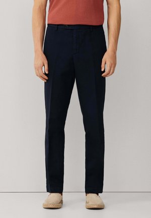 Man draagt slim fit donkerblauwe broek, een roestkleurig T-shirt met korte mouwen en beige espadrilles, staand tegen een effen achtergrond.