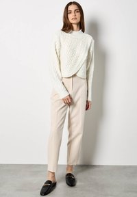 Maglione color crema a trecce con collo alto, abbinato a pantaloni beige su misura e mocassini neri con dettagli dorati.