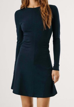 Femme portant une robe noire cintrée à manches longues avec une légère évasement et un col rond, debout devant un fond clair uni.